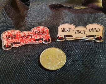 May include: Two decorative pins with the Latin phrase "Mors Vincit Omnia" (Death conquers all) on a scroll. One pin is white with black lettering and the other is red with white lettering.