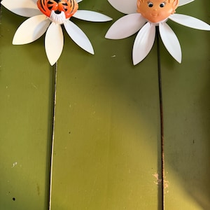 以下が含まれることがあります： 白いプラスチック製のデイジーの花びらを持つ2つのガーデンスピナー。片方の中央には成形されたオレンジと黒のトラの顔があり、もう片方にはピーチトーンの妖精の顔が付いています。どちらも細く風化した金属製の支柱に取り付けられています。