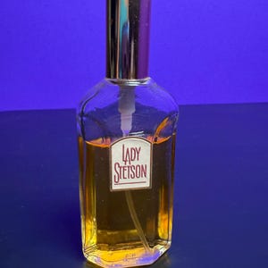 May include: A vintage perfume bottle with a gold-coloured cap and a white pump. The clear glass bottle contains a golden liquid and features a label reading "LADY STETSON" in red. The background is a deep blue, highlighting the bottle's design.