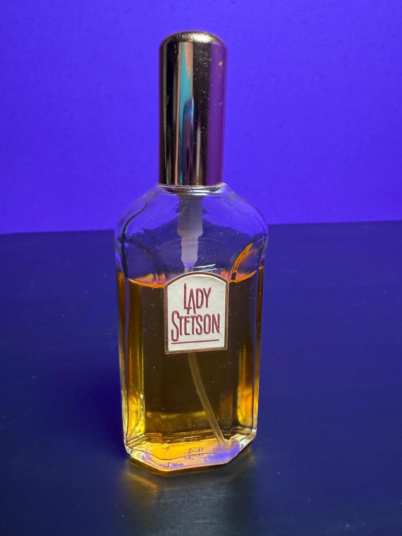 May include: A vintage perfume bottle with a gold-coloured cap and a white pump. The clear glass bottle contains a golden liquid and features a label reading "LADY STETSON" in red. The background is a deep blue, highlighting the bottle's design.