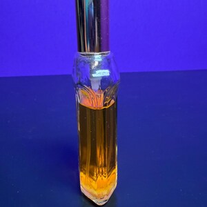 May include: A clear glass perfume bottle with a gold-coloured cap and a rectangular shape. The bottle is filled with a golden-orange liquid. The bottle has a decorative design on the front and is set against a blue background.