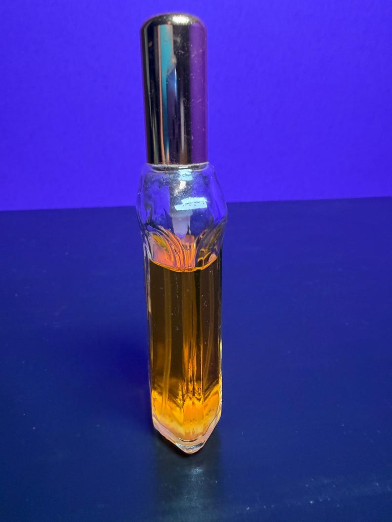 May include: A clear glass perfume bottle with a gold-coloured cap and a rectangular shape. The bottle is filled with a golden-orange liquid. The bottle has a decorative design on the front and is set against a blue background.