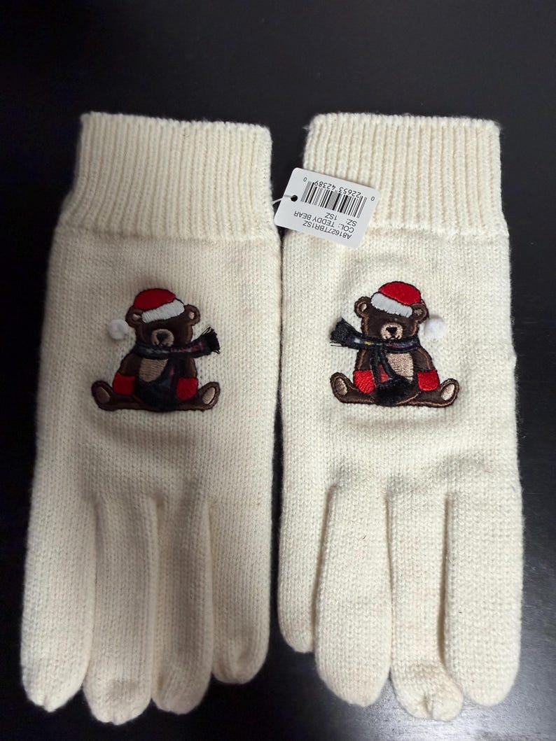 May include: A pair of ivory-colored knit gloves. Each glove features an embroidered teddy bear wearing a red Santa hat, a black scarf, and red boots. The gloves have a ribbed cuff and are designed for winter wear.