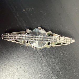 K&ouml;nnte beinhalten: Eine silberfarbene Armbanduhr mit rechteckigem Gliederarmband. Das Zifferblatt der Uhr ist rund mit einer silbernen L&uuml;nette. Die R&uuml;ckseite der Uhr ist sichtbar, mit dem Text "Steel Back". Die Uhr ist vor einem dunklen Hintergrund platziert.