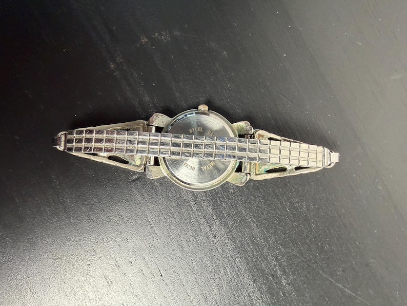 K&ouml;nnte beinhalten: Eine silberfarbene Armbanduhr mit rechteckigem Gliederarmband. Das Zifferblatt der Uhr ist rund mit einer silbernen L&uuml;nette. Die R&uuml;ckseite der Uhr ist sichtbar, mit dem Text "Steel Back". Die Uhr ist vor einem dunklen Hintergrund platziert.