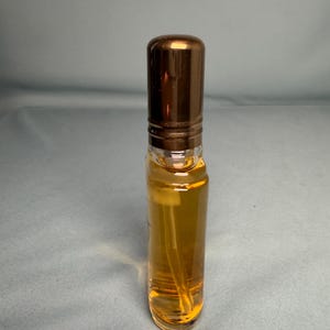 May include: A clear glass perfume bottle with a bronze-colored cap. The bottle contains a golden-colored liquid and a white spray nozzle. The bottle is cylindrical and has a simple, elegant design.
