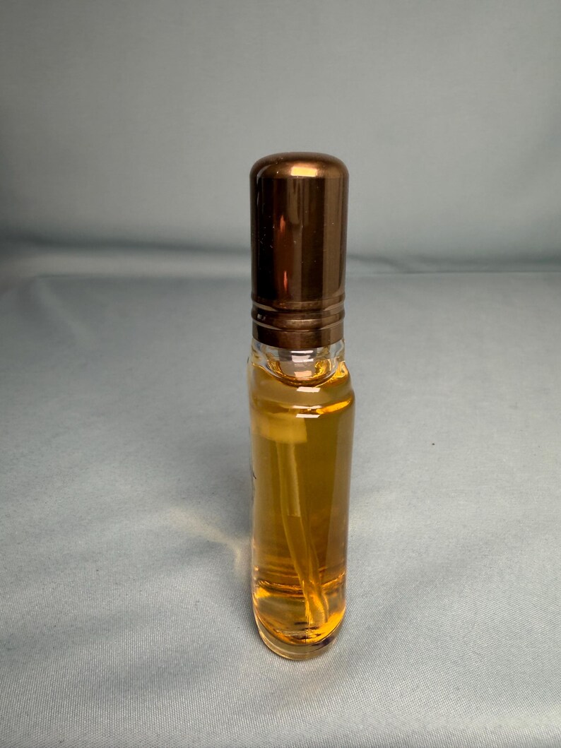 May include: A clear glass perfume bottle with a bronze-colored cap. The bottle contains a golden-colored liquid and a white spray nozzle. The bottle is cylindrical and has a simple, elegant design.