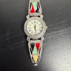 K&ouml;nnte beinhalten: Eine silberfarbene Quarzuhr mit cremefarbenem Zifferblatt und r&ouml;mischen Ziffern. Das Uhrenarmband weist farbenfrohe geometrische Designs in Rot, Schwarz, Gr&uuml;n und Gelb auf. Die Uhr wird auf einer dunklen Oberfl&auml;che pr&auml;sentiert.