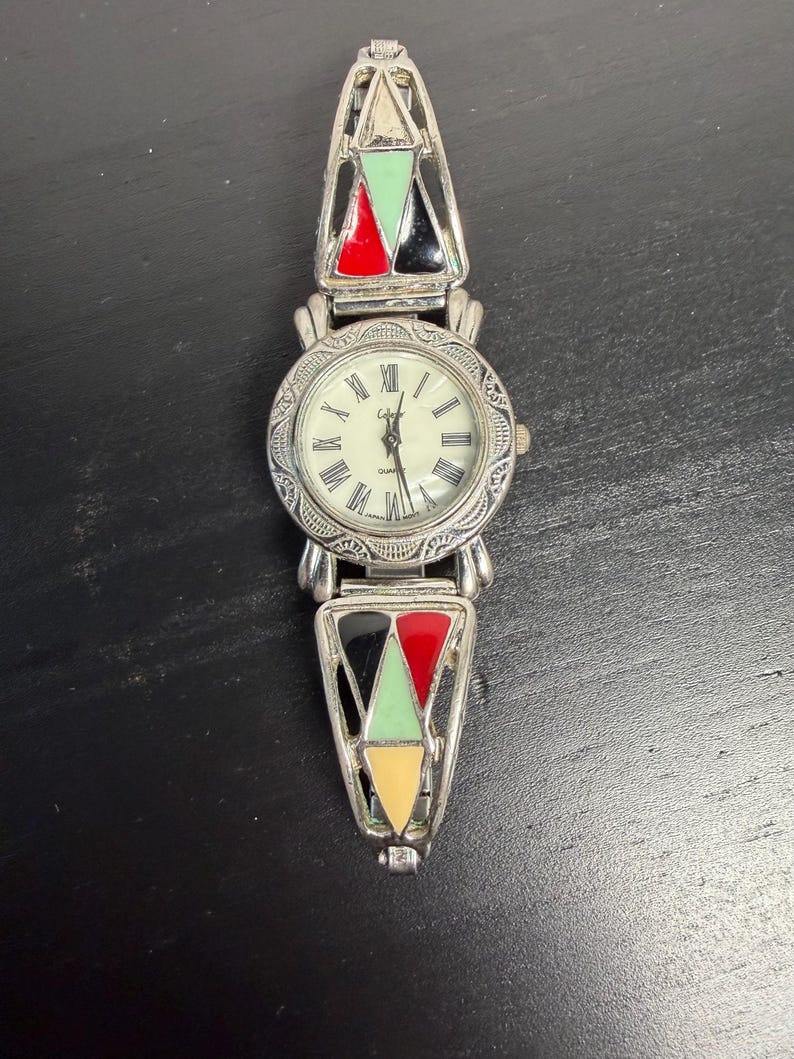 K&ouml;nnte beinhalten: Eine silberfarbene Quarzuhr mit cremefarbenem Zifferblatt und r&ouml;mischen Ziffern. Das Uhrenarmband weist farbenfrohe geometrische Designs in Rot, Schwarz, Gr&uuml;n und Gelb auf. Die Uhr wird auf einer dunklen Oberfl&auml;che pr&auml;sentiert.