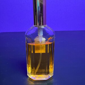 May include: A clear glass perfume bottle with a gold-coloured cap and a label. The bottle contains a golden-coloured liquid. The bottle is on a blue surface.