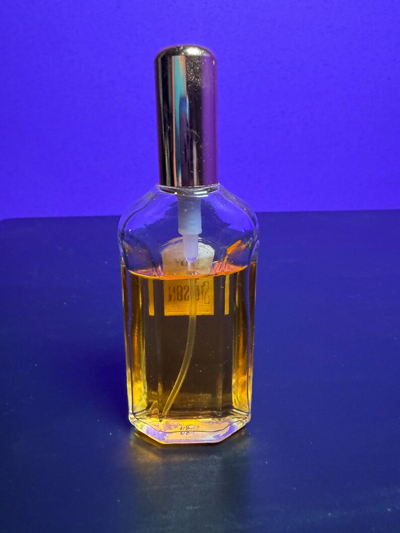 May include: A clear glass perfume bottle with a gold-coloured cap and a label. The bottle contains a golden-coloured liquid. The bottle is on a blue surface.