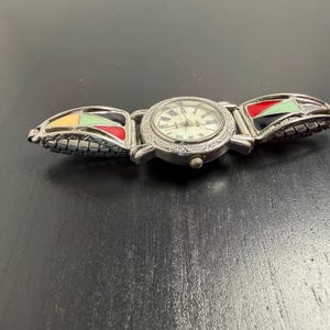 K&ouml;nnte beinhalten: Silberfarbene Armbanduhr mit rundem Zifferblatt und r&ouml;mischen Ziffern. Das Armband der Uhr hat farbenfrohe geometrische Designs in Gelb, Gr&uuml;n, Rot und Schwarz. Die Uhr liegt auf einer dunklen Oberfl&auml;che.