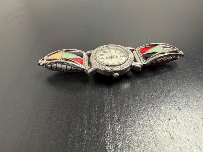 K&ouml;nnte beinhalten: Silberfarbene Armbanduhr mit rundem Zifferblatt und r&ouml;mischen Ziffern. Das Armband der Uhr hat farbenfrohe geometrische Designs in Gelb, Gr&uuml;n, Rot und Schwarz. Die Uhr liegt auf einer dunklen Oberfl&auml;che.