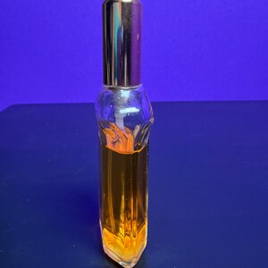 May include: A small, clear glass perfume bottle with a gold-coloured cap. The bottle is filled with a golden-orange liquid. The bottle has a unique, geometric shape and is set against a blue background.