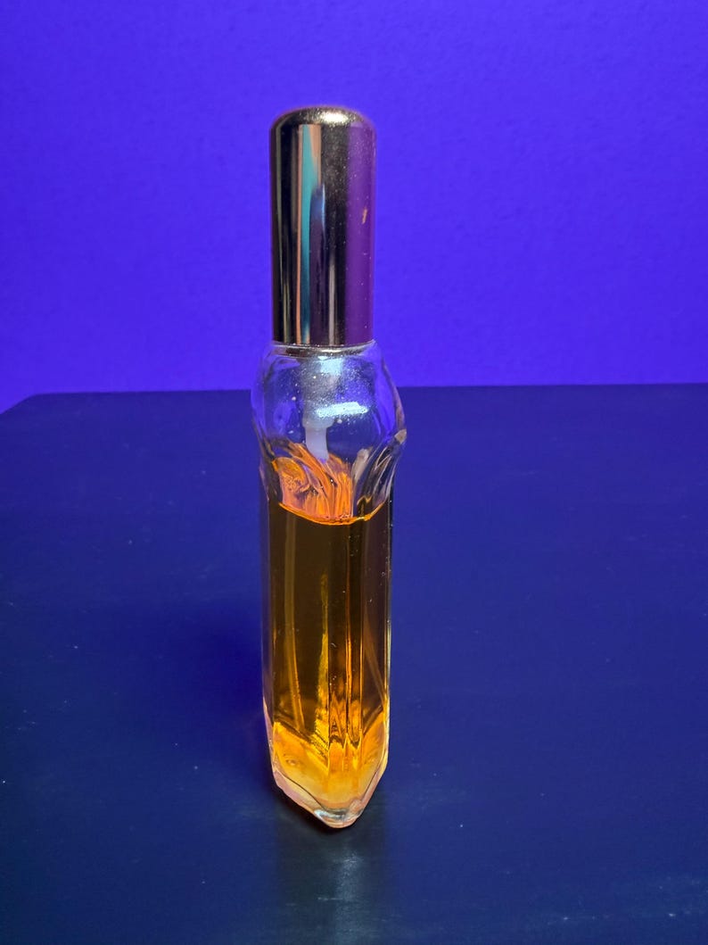 May include: A small, clear glass perfume bottle with a gold-coloured cap. The bottle is filled with a golden-orange liquid. The bottle has a unique, geometric shape and is set against a blue background.