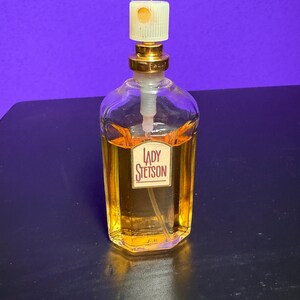 May include: A clear glass perfume bottle with a gold-coloured cap and a label that reads "LADY Stetson." The bottle contains a golden-coloured liquid. The background is a solid purple colour.