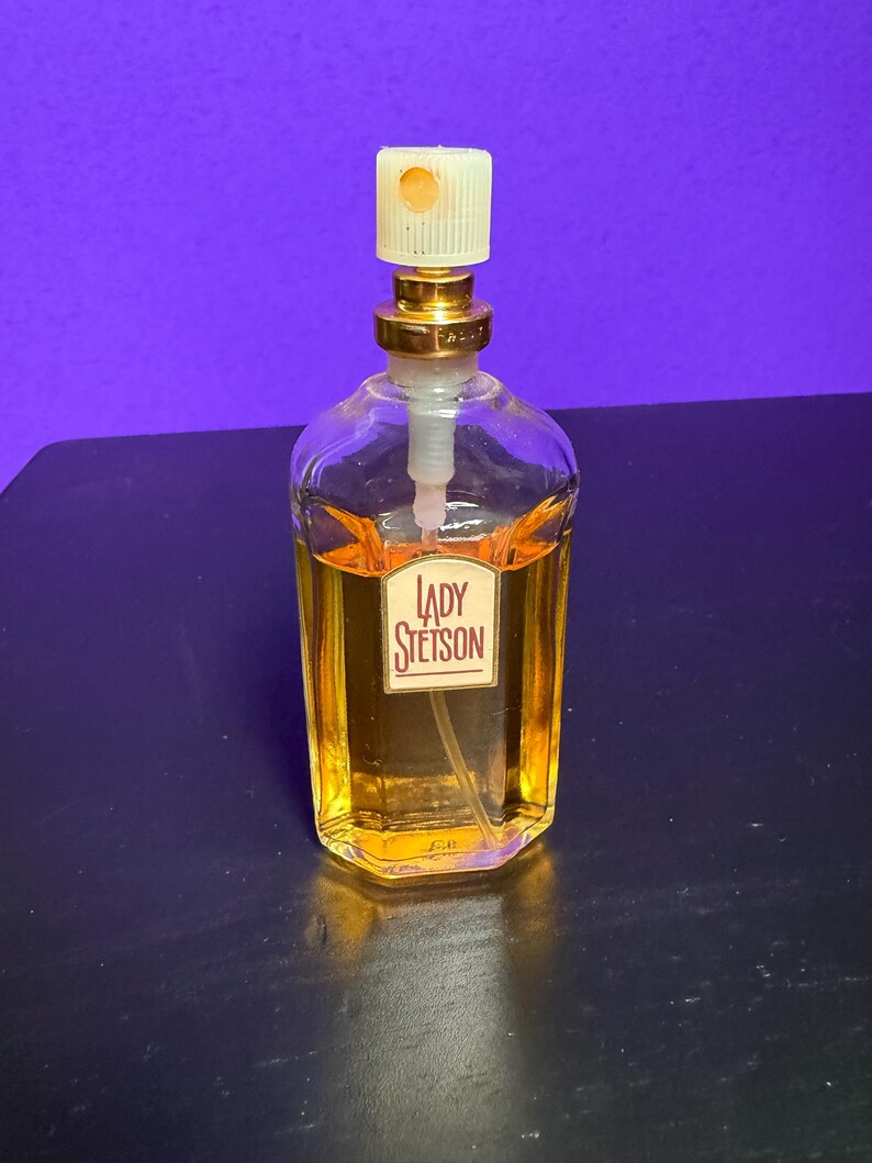 May include: A clear glass perfume bottle with a gold-coloured cap and a label that reads "LADY Stetson." The bottle contains a golden-coloured liquid. The background is a solid purple colour.