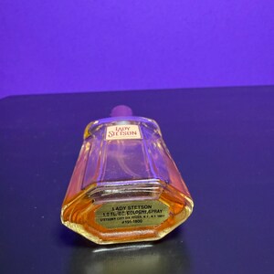 May include: A vintage Lady Stetson cologne spray bottle. The clear glass bottle has a trapezoidal shape with a purple cap. The label reads "Lady Stetson 1.0 fl. oz. Cologne Spray". The liquid inside is amber-coloured.