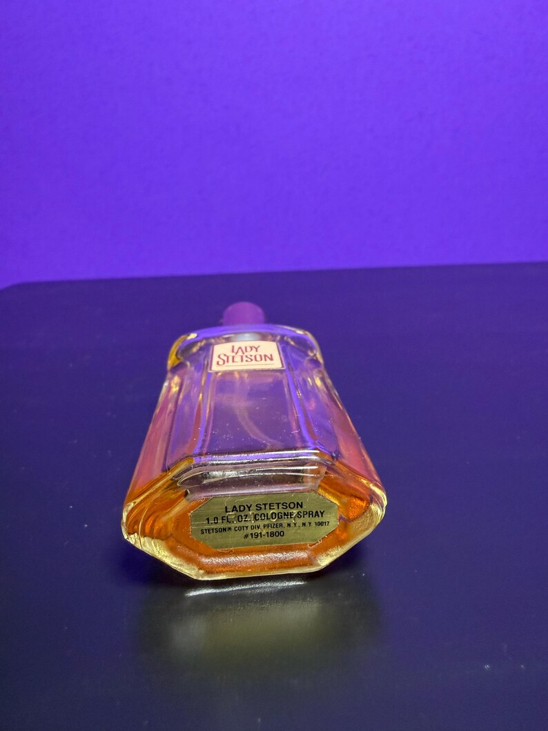May include: A vintage Lady Stetson cologne spray bottle. The clear glass bottle has a trapezoidal shape with a purple cap. The label reads "Lady Stetson 1.0 fl. oz. Cologne Spray". The liquid inside is amber-coloured.