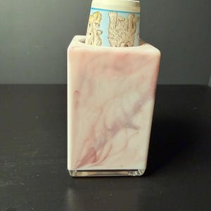 May include: A rectangular, pink and white marbled resin holder with a smooth, glossy finish. A cylindrical object with a light blue border and a printed illustration of a tan, furry creature is inserted into the top opening of the holder.