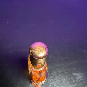 May include: A small, clear glass perfume bottle with an orange liquid inside. The bottle has a gold-coloured cap. The bottle is on a dark surface with a purple background.