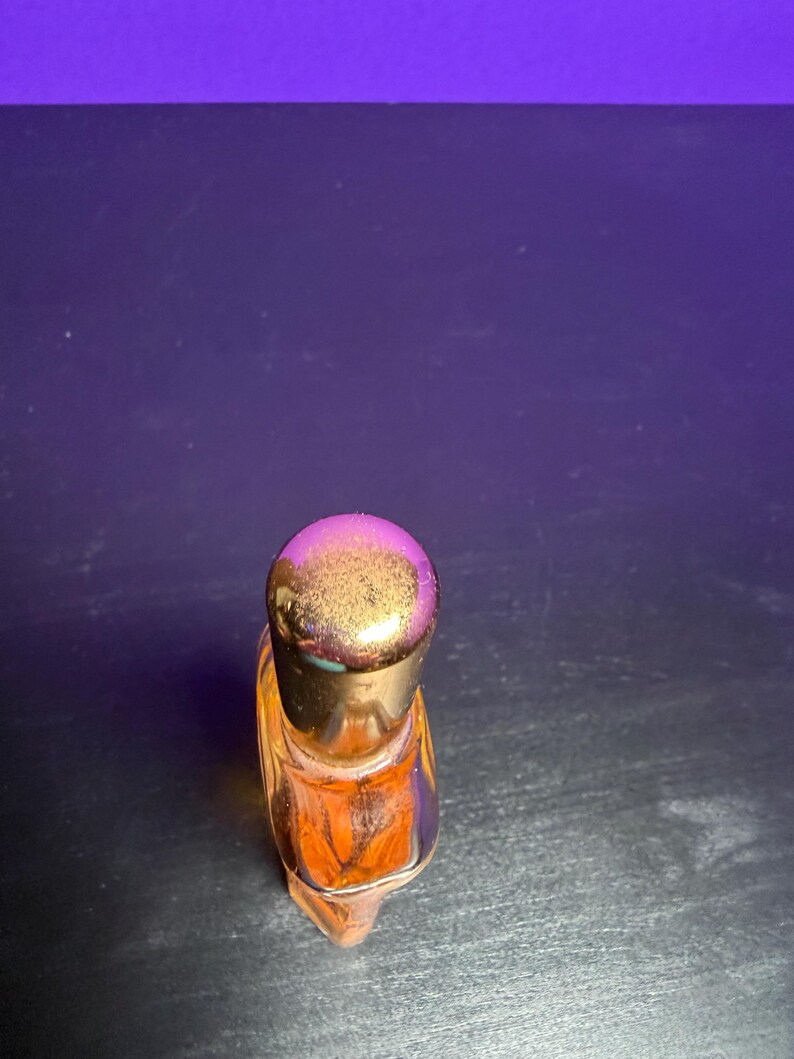 May include: A small, clear glass perfume bottle with an orange liquid inside. The bottle has a gold-coloured cap. The bottle is on a dark surface with a purple background.