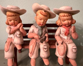 May include: Three ceramic figurines of children in cowboy hats and outfits seated on a wooden bench. The figurines are painted in soft pinks, whites, and browns. Each child has a different accessory, including a scarf and a shield.