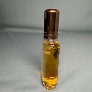 May include: A small, clear glass perfume bottle filled with a golden liquid. The bottle has a rounded rectangular shape and a shiny, bronze-colored cap. The perfume bottle is standing upright.