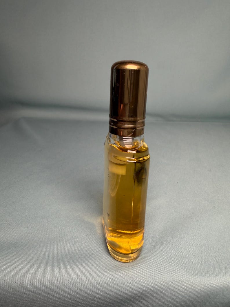 May include: A small, clear glass perfume bottle filled with a golden liquid. The bottle has a rounded rectangular shape and a shiny, bronze-colored cap. The perfume bottle is standing upright.