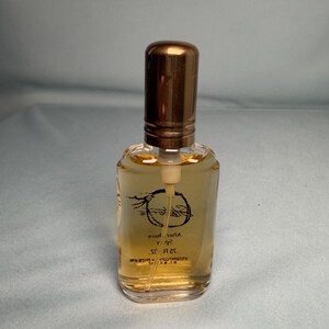 May include: A small, clear glass perfume bottle with a bronze-colored cap. The bottle contains a light amber liquid. The label features cursive text and a circular design. The bottle is rectangular with rounded edges.