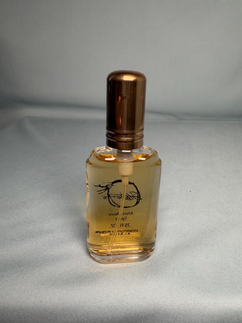 May include: A small, clear glass perfume bottle with a bronze-colored cap. The bottle contains a light amber liquid. The label features cursive text and a circular design. The bottle is rectangular with rounded edges.