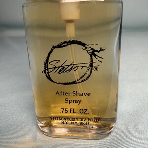 May include: A clear glass bottle of Stetson After Shave Spray. The bottle has a rectangular shape with rounded edges and a beige liquid inside. The label features the Stetson logo and product information, including the volume of .75 FL. OZ.