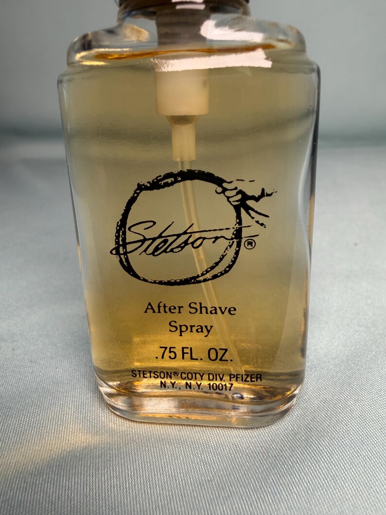 May include: A clear glass bottle of Stetson After Shave Spray. The bottle has a rectangular shape with rounded edges and a beige liquid inside. The label features the Stetson logo and product information, including the volume of .75 FL. OZ.