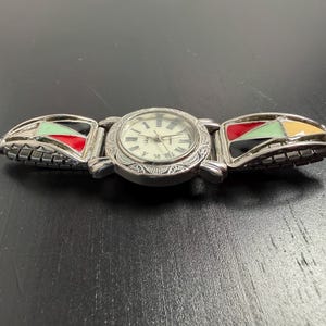 K&ouml;nnte beinhalten: Silberfarbene Armbanduhr mit cremefarbenem Zifferblatt und r&ouml;mischen Ziffern. Das Armband hat farbenfrohe geometrische Designs in Rot, Gr&uuml;n, Schwarz und Gelb. Die Uhr liegt auf einer dunklen Oberfl&auml;che.