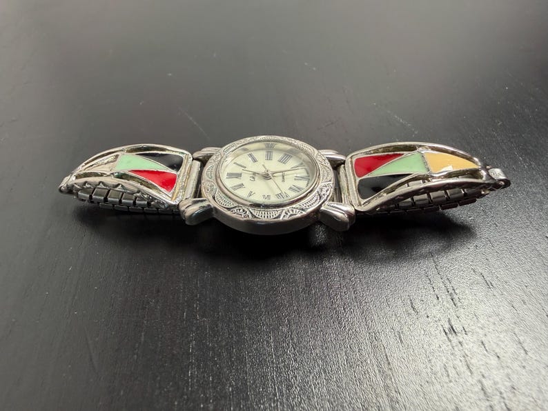 K&ouml;nnte beinhalten: Silberfarbene Armbanduhr mit cremefarbenem Zifferblatt und r&ouml;mischen Ziffern. Das Armband hat farbenfrohe geometrische Designs in Rot, Gr&uuml;n, Schwarz und Gelb. Die Uhr liegt auf einer dunklen Oberfl&auml;che.
