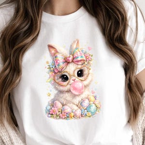 Puede incluir: Camiseta blanca con una ilustración de acuarela de un conejito con gafas y un lazo, soplando una burbuja de chicle. El conejito está rodeado de flores y huevos de Pascua. La camiseta es de material suave.