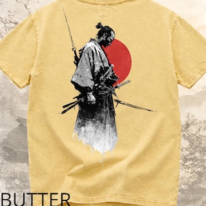 Puede incluir: Camiseta de color mantequilla con un diseño de un guerrero samurái, espadas y un sol rojo. El diseño es en blanco y negro, con la palabra "BUTTER" impresa en la parte inferior. La camiseta tiene un aspecto vintage y desgastado.