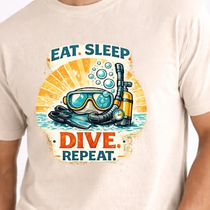 Puede incluir: Camiseta beige con un gráfico colorido de equipo de buceo, que incluye una máscara, un esnórquel y aletas, sobre un fondo de sol. El texto "EAT. SLEEP. DIVE. REPEAT." se muestra arriba y abajo del gráfico.