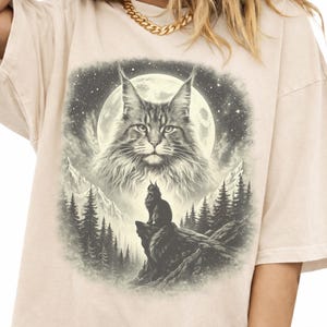Puede incluir: Camiseta beige con un gráfico detallado de un gato Maine Coon frente a una luna llena, montañas y una silueta de gato sobre una roca. El diseño utiliza tonos grises y negros.