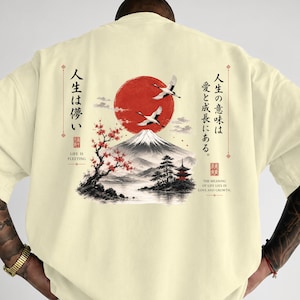 Puede incluir: Una camiseta de color crema con un gráfico en la espalda que muestra un sol rojo, el monte Fuji, dos grullas volando, un cerezo en flor y una pagoda. Texto en japonés y las frases en inglés "Life is fleeting" y "The meaning of life lies in love and growth" están impresas a los lados.