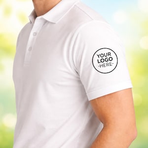Puede incluir: Polo blanco con tapeta de tres botones y mangas cortas. Un círculo negro con el texto "YOUR LOGO -HERE-" está impreso en la manga izquierda. La camisa está hecha de un material liso, probablemente algodón.