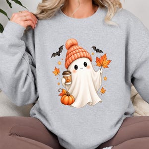 Puede incluir: Una sudadera gris claro de cuello redondo con un gráfico central de un fantasma blanco con un gorro de punto naranja. El fantasma sostiene una taza de café y una hoja de arce naranja, rodeado por una calabaza, dos murciélagos negros y estrellas.