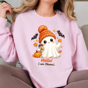 Puede incluir: Una sudadera rosa claro con un gráfico de un fantasma blanco con un gorro de punto naranja, sosteniendo una taza de café y junto a una pequeña calabaza naranja. El diseño incluye murciélagos, hojas de otoño y el texto Hello! I am (Name).