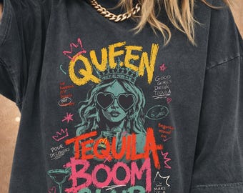 Può includere: T-shirt grigio scuro con un design grafico. Il design presenta la parola "QUEEN" in giallo, una donna con occhiali da sole a forma di cuore e le parole "TEQUILA BOOM CLUB" in rosa e rosso. Il testo aggiuntivo include "Good Girls Drink Tequila."
