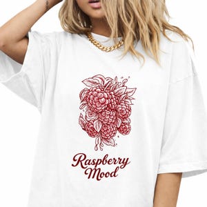 Puede incluir: Camiseta blanca con una ilustración de frambuesas rojas, hojas y gotas de agua. El texto "Raspberry Mood" está escrito en cursiva debajo. La camiseta tiene un corte holgado.