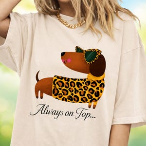 Puede incluir: Camiseta beige con un perro salchicha de dibujos animados que lleva gafas de sol con estampado de leopardo y un suéter a juego. El perro salchicha tiene la nariz rosa y está sonriendo. El texto "Always on Top..." está escrito en cursiva debajo.