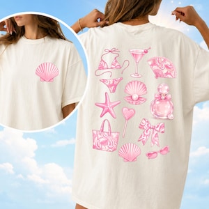 Puede incluir: Una camiseta color crema con un pequeño gráfico de concha rosa en el pecho izquierdo. La parte trasera muestra una colección de ilustraciones rosas que incluyen un bikini, copa de martini, abanico, estrella de mar, perla en concha, frasco de perfume, piruleta de corazón, bolso, lazo y gafas de sol.