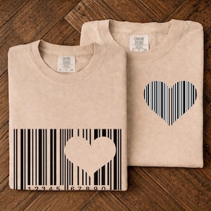 Puede incluir: Dos camisetas beige con diseños de corazón. Una camiseta presenta un diseño de código de barras con un recorte de corazón, y la otra tiene un corazón hecho de líneas negras verticales. Las camisetas están sobre una superficie de madera.
