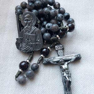 Puede incluir: Un rosario con cuentas grises oscuras y negras, un crucifijo plateado y una medalla de santo plateada. Las cuentas están ensartadas en un cordón verde. El crucifijo presenta a Jesucristo.