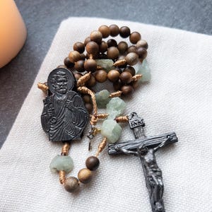 May include: A rosary with wooden beads, light green stone accents, and a dark metal crucifix. The rosary features a detailed pendant of a religious figure and is displayed on a white cloth.